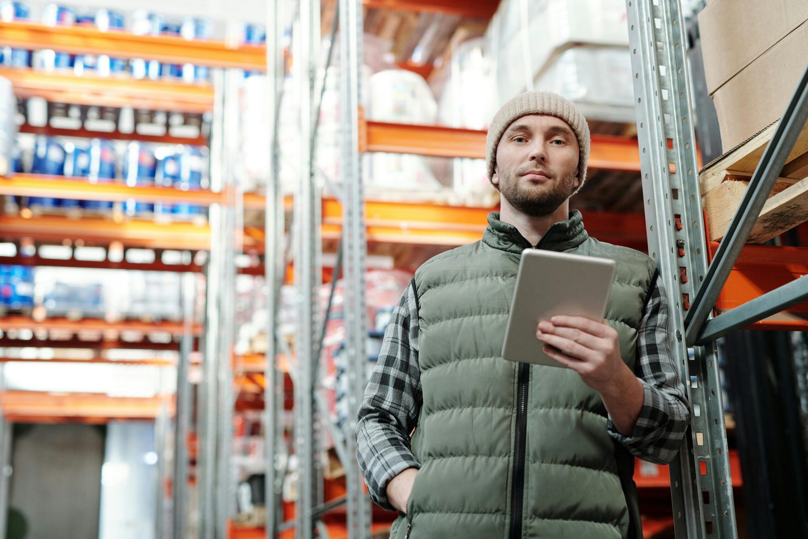 Young male warehouse manager in a beanie holding a tablet in a storeroom.